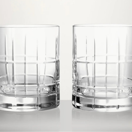 Two clear cut-crystal whiskey glasses with vertical fluting and a thick base, shown side by side on a light gray reflective surface against a light gray background.