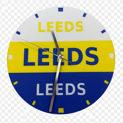 A circular wall clock featuring the Leeds club colors, with a white top half, yellow middle band, and blue bottom half, all printed with the word 'LEEDS'. The clock has silver hands and small blue and white hour markers.