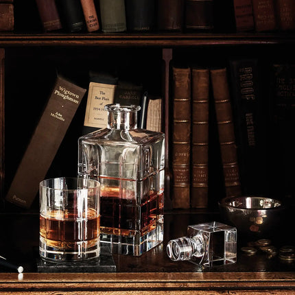 A set of clear glass decanters and a matching whiskey glass with amber liquor, arranged on a dark wooden table in front of a bookshelf filled with vintage books.