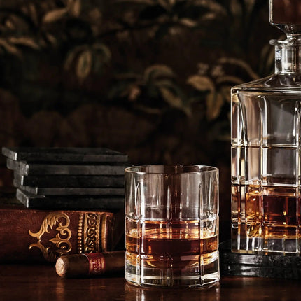 A clear whiskey tumbler glass and a matching clear decanter filled with amber liquor, styled on a dark wooden surface with a cigar, stacked dark glasses, and an ornate book against a dark patterned background.