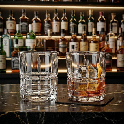 Two clear cut-glass rocks tumblers on a dark bar counter, with one empty and one filled with amber liquor and an orange peel garnish, in front of a softly blurred bar shelf of bottles.