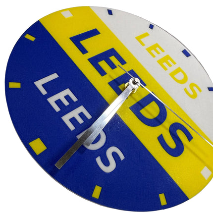 A circular wall clock featuring the Leeds United football club colors and branding, with blue, yellow, and white diagonal sections and the word 'LEEDS' printed across the face. The clock is shown mounted on a light gray wall.