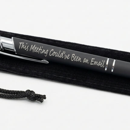 A black and silver ballpoint pen with the text 'This Meeting Could've Been an Email' printed on it, resting on a black fabric pouch.