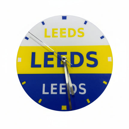 A circular wall clock featuring the Leeds United football club colors, with a white top half, yellow middle band, and blue bottom half, all printed with the word 'LEEDS'. The clock has silver hands and simple colored hour markers.