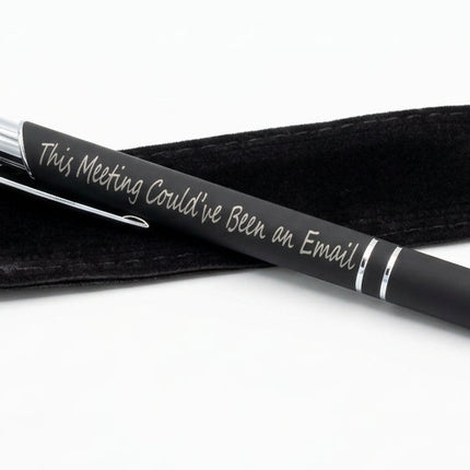 A black ballpoint pen with silver lettering that reads 'This Meeting Could've Been an Email!' resting on a black guitar headstock, with a black drawstring pouch beside it.