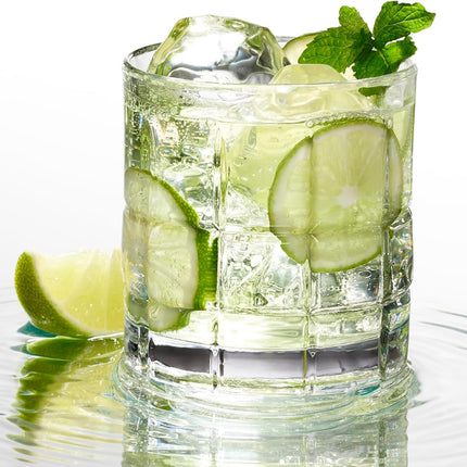 A clear textured glass filled with a light green drink, ice cubes, lime slices, and mint leaves, set on a white reflective surface with a lime wedge beside it.