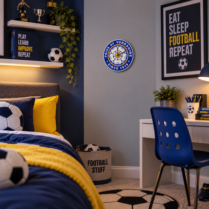 A styled boy's bedroom featuring a blue and yellow color scheme with soccer-themed decor, including a Leicester City FC crest on the wall, soccer pillows, a desk with a blue chair, and framed motivational posters.