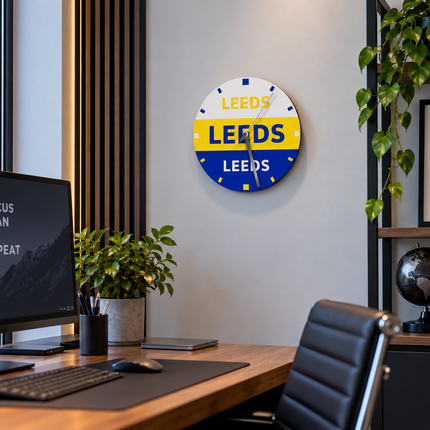 A round wall clock with 'LEEDS' repeated in white, yellow, and blue segments, mounted on a light gray wall in a modern home office setting with a desk, computer monitor, and plants.