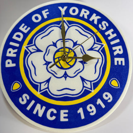 A round wall clock featuring the blue, white, and yellow crest of Pride of Yorkshire, with white hands and no traditional hour markers.