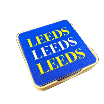 A square blue coaster with a white border, featuring the word 'LEEDS' printed three times in alternating yellow and white block letters, set against a dark brown background.
