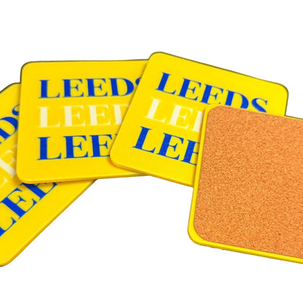 Four square yellow coasters on a dark surface, with three showing blue 'LEEDS' text and one showing a brown cork backing with a yellow border.