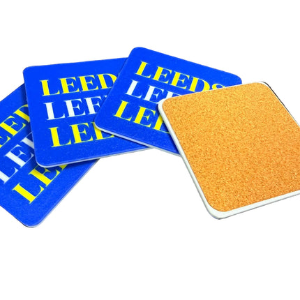Four square coasters on a dark surface, including three blue coasters with yellow and white 'LEED' text and one tan cork coaster with a white border.