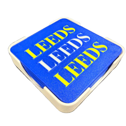 A stack of square blue coasters with a white plastic frame, featuring the word 'LEEDS' printed in alternating yellow and white block letters, set against a dark background.