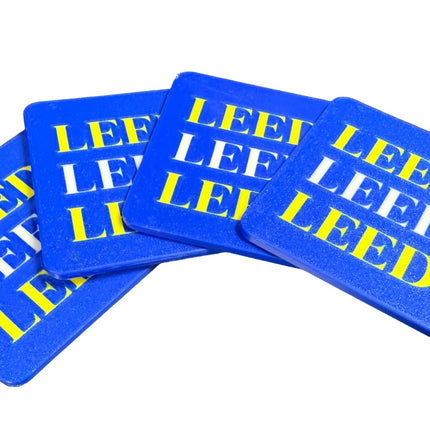 Four blue square coasters with rounded corners, stacked slightly offset on a dark surface. Each coaster features the text 'LEE LEEDS' printed in alternating white and yellow block letters.