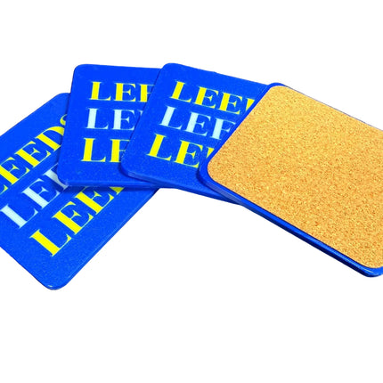 Four square coasters on a dark surface, with three showing blue tops printed with repeated yellow and white 'LEE' lettering and one showing a tan cork-like underside with a blue border.