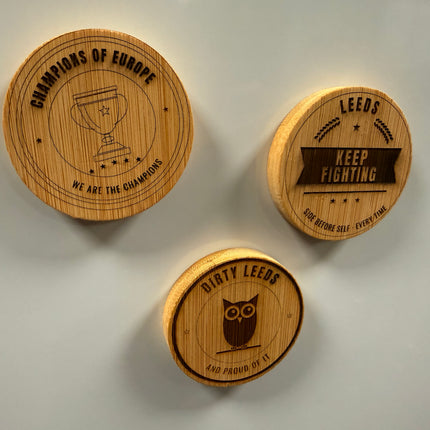 Three round wooden magnets with engraved sports designs and text, displayed on a light gray surface.