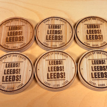 Six round wooden poker chips engraved with 'LEEDS LEEDS LEEDS!' are arranged on a light wood tabletop, with stacks of similar chips and black circular game pieces in the background.