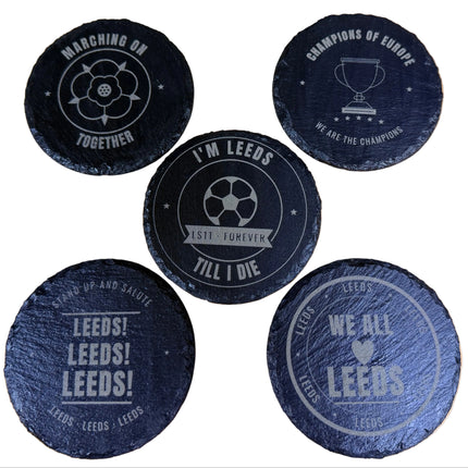 Five round black slate coasters with engraved white football-themed designs and text, arranged on a light wood surface.