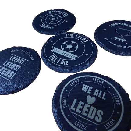 Five dark slate coasters with Leeds United football club designs and slogans, arranged on a light wood tabletop.