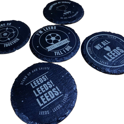Five round black slate coasters with white printed designs and text, including 'LEEDS' and soccer-themed graphics, arranged on a light wood tabletop.