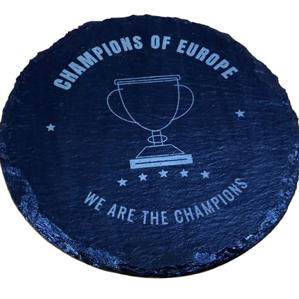 A round black slate coaster with a white printed trophy graphic and the text 'CHAMPIONS OF EUROPE' and 'WE ARE THE CHAMPIONS', placed on a light wood surface.