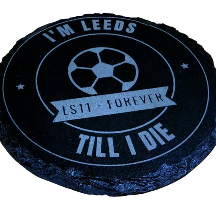 A round black slate coaster with a light blue printed design featuring a soccer ball and the text 'I'M LEEDS LS11 FOREVER TILL I DIE', displayed on a light wood background.