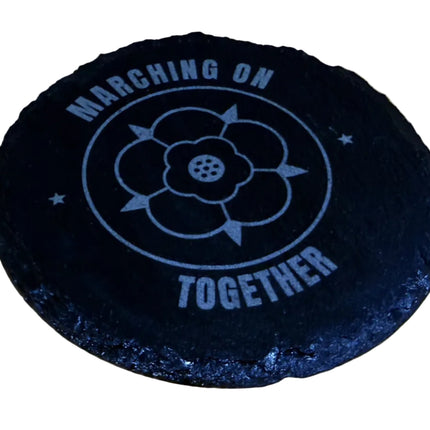 A round black coaster with a white printed design reading 'MARCHING ON TOGETHER' and a flower emblem, placed on a light wood surface.