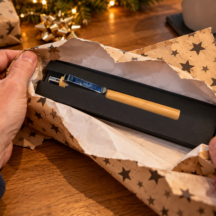 A close-up of hands unwrapping brown star-patterned gift wrapping paper from a black presentation box on a wooden table. Inside the box is a slim wooden-handled tool or accessory with a metallic blue and gold section.