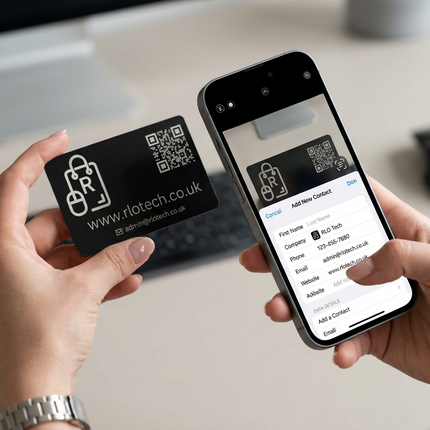 Two hands holding a black contact card and a smartphone showing the mobile app that creates and manages the contact card, set against a blurred office desk background.