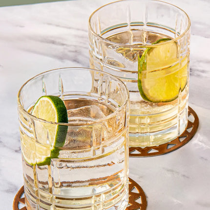 Two clear textured glass tumblers filled with a pale golden drink and garnished with lime slices, resting on decorative round coasters on a white marble surface.