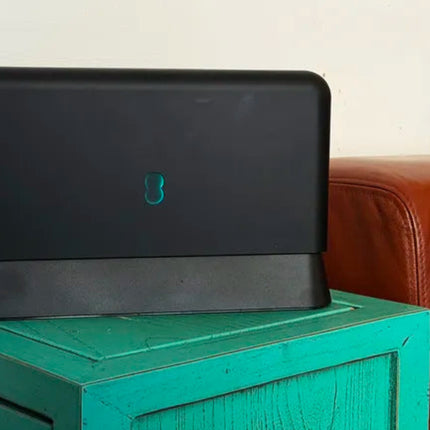 A black rectangular electronic device with a small teal logo, resting on a distressed teal wooden box next to a brown leather sofa against a plain off-white wall.